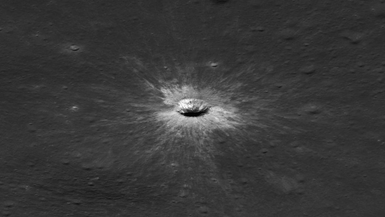 A fresh impact crater on the gray lunar surface, with bright rays spreading outward.